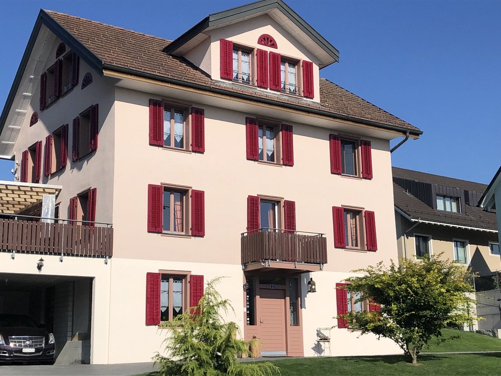 Mehrstöckiges Haus mit weißen Wänden, roten Fensterläden, einem Balkon und einem kleinen Baum im Vorgarten, blauer Himmel.