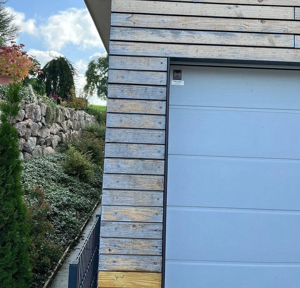 Ansicht eines Hauses mit Holzverkleidung und einer Garage, im Hintergrund ein Garten mit Pflanzen und Steinen, bei sonnigem Himmel.