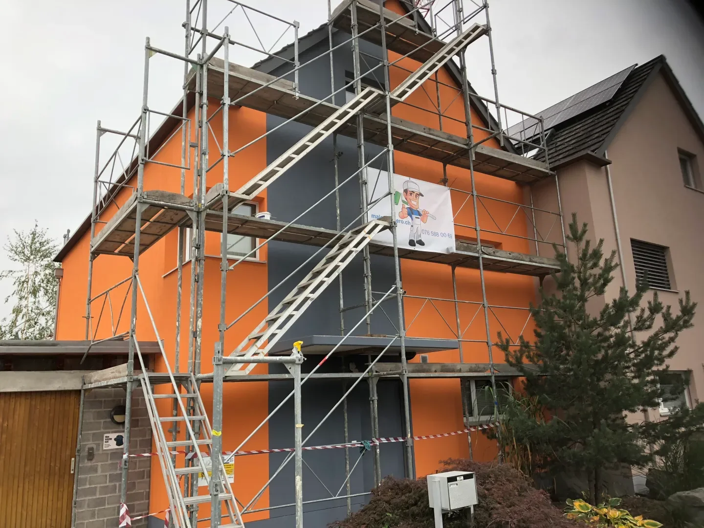 Ein mehrstöckiges Gebäude wird mit Gerüsten und Baugerüstmaterialien renoviert oder gestrichen. Es gibt eine Leiter, die an die Gerüste angebracht ist, und eine Baustellenabsperrung. Eine Plane mit einer humorvollen Illustration eines Handwerkers hän