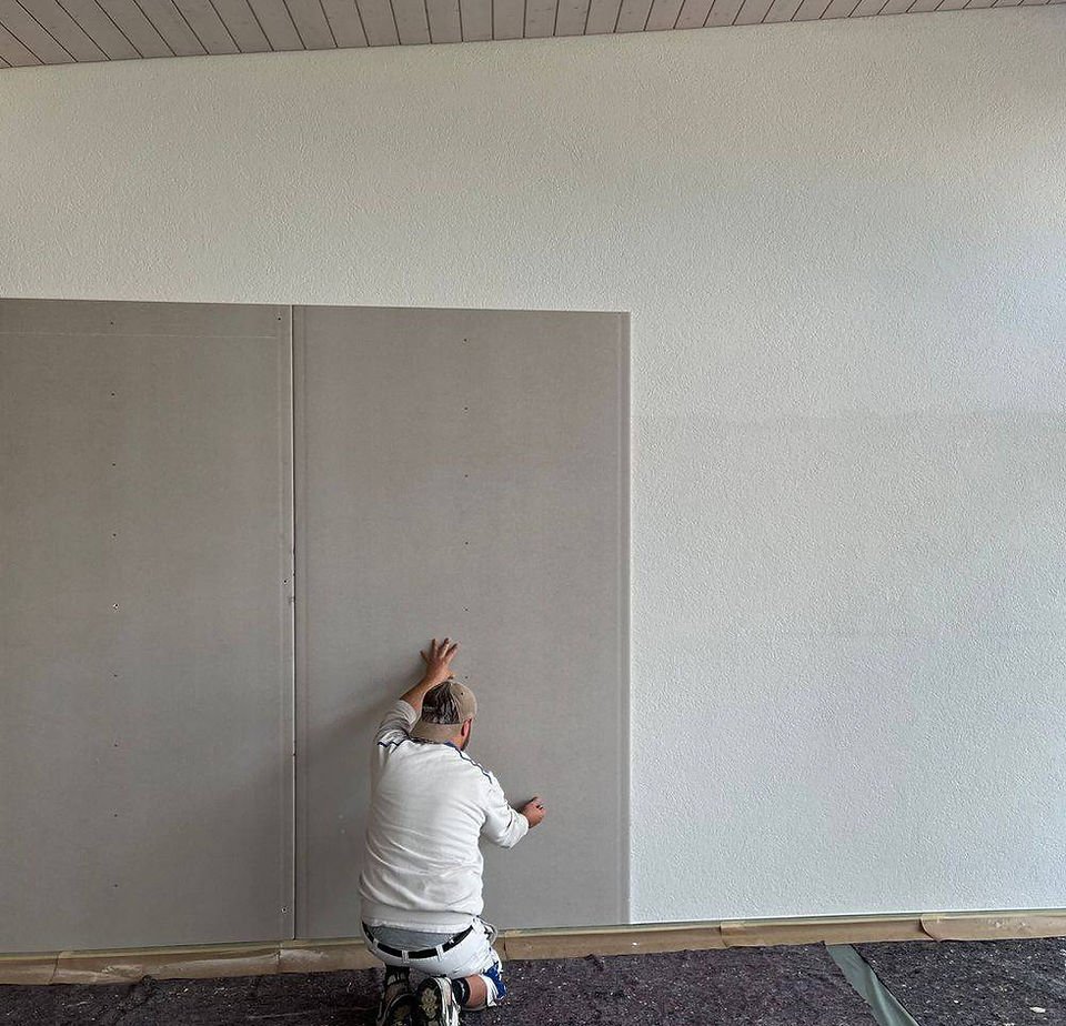 Ein Maler kniet auf dem Boden und versucht, eine zweite Wandplatte zu befestigen.