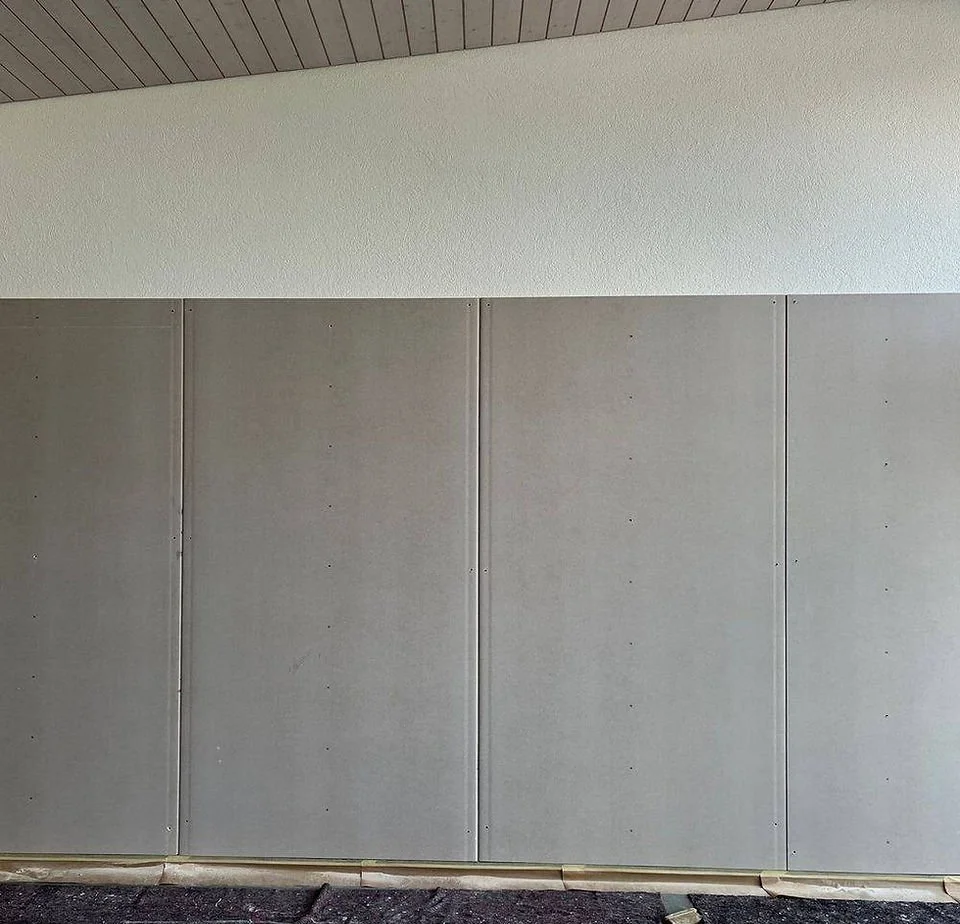 Vier graue Wandpaneele auf einer Wand mit einer weißen Wand oben, der Boden ist dunkel und uneben.