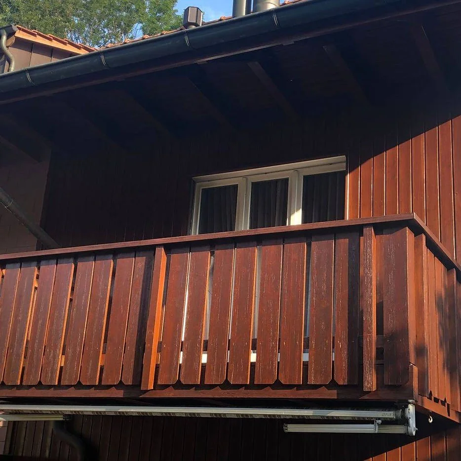 Ansicht eines Holzhäuschens mit einer kleinen Balkonveranda und einem Fenster, das von innen mit Vorhängen bedeckt ist.