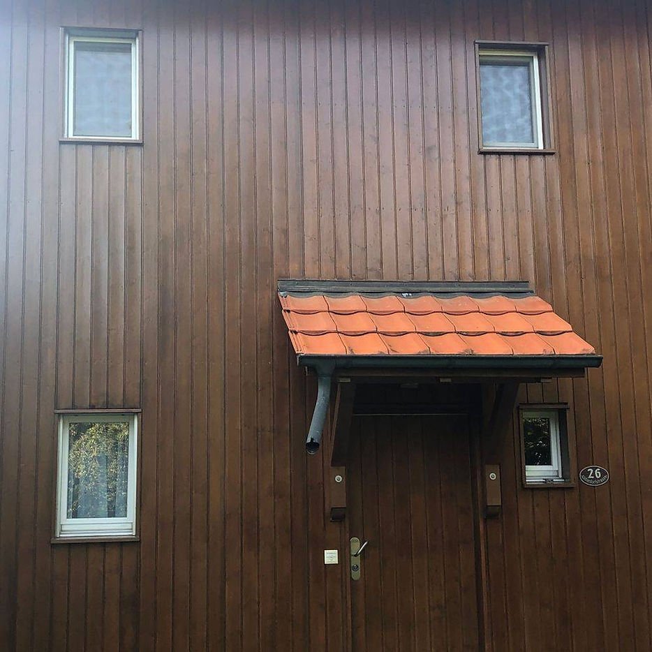 Fassade eines Hauses mit vertikalen Holzwänden, vier kleine Fenster und einem überdachten Eingang mit rotem Ziegeldach.
