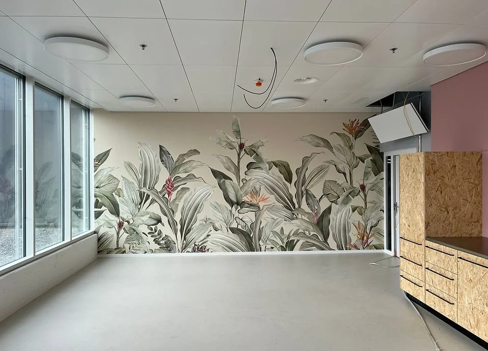 Ein leerer Raum mit einer Wand, die mit einer botanischen Wandmalerei mit großen Blättern und Blumen dekoriert ist, große Fenster an der Seite, eine rustikale Holz-Schreibtisch oder -Schrank an der rechten Ecke.