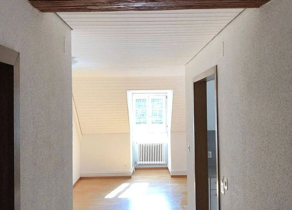 Leeres Zimmer mit Dachschräge, Fenster und Holzboden in einem hell erleuchteten Raum.