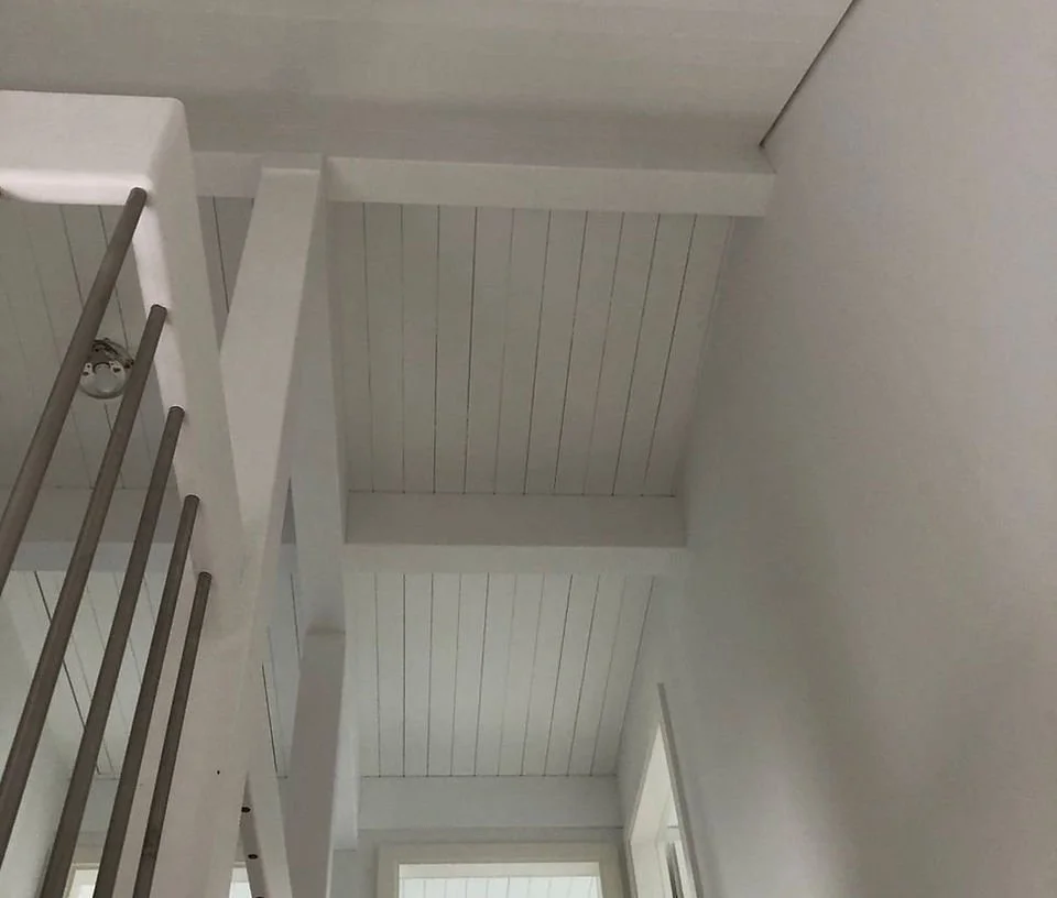 Innenraum mit weiß gestrichenen Wänden und einer Decke mit sichtbaren Holzbalken, daneben ein Fenster und ein Holzgeländer.