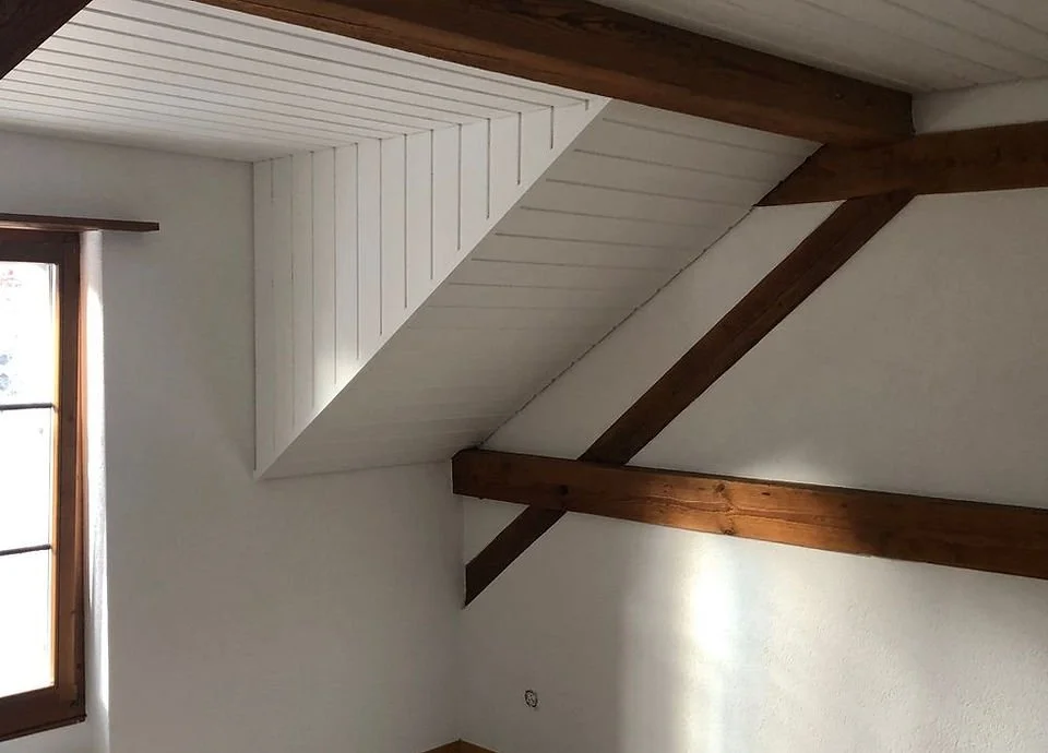 Innenraum eines Dachgeschosses mit weiß gestrichener Wand und sichtbaren Holzbalken, eine kleine Fensteröffnung lässt Tageslicht herein.