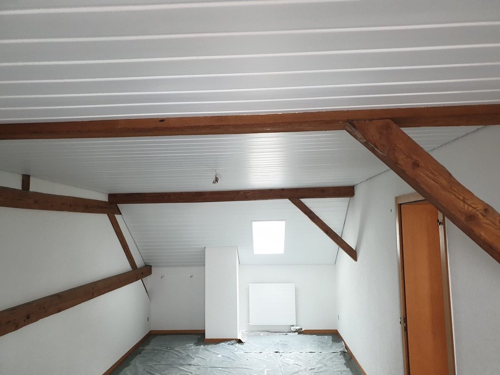 Innenraum eines Dachbodens mit weißen Wänden, Holzbalken und einem schrägen weißen Deckenpaneel. Es liegt eine Plane auf dem Boden und an der Wand hängt eine Heizkörper.