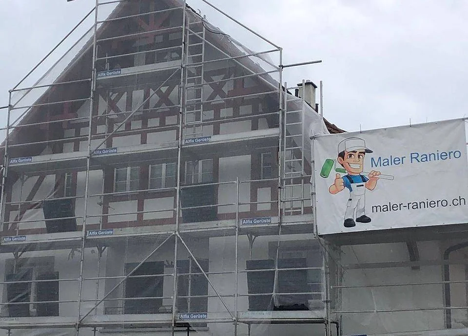 Fassade eines Hauses im Renovierungsprozess, umgeben von Gerüsten, mit einem Banner eines Malers mit Pinsel und Farbrolle, auf dem steht "Maler Raniero" und die Webseite "maler-raniero.ch".