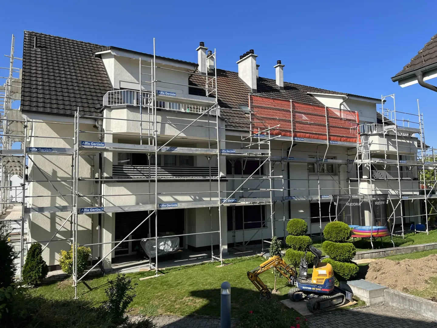Mehrstöckiges Mehrfamilienhaus unter Renovierung mit Baugerüsten, Garten mit bepflanzten Büsche, Spielzeugpools, Handwerksmaschinen und Bauarbeiten im Vordergrund, blauer Himmel und Sonnenschein.