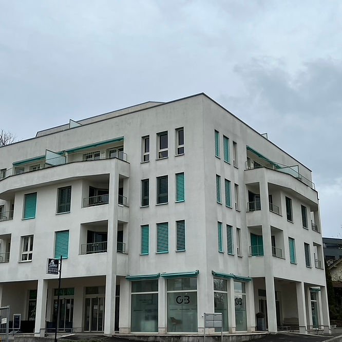 Mehrstöckiges weißes Gebäude mit mehreren Balkonen und großen Fenstern, einige Fenster mit grünen Rollläden, Geschäftsladen im Erdgeschoss.