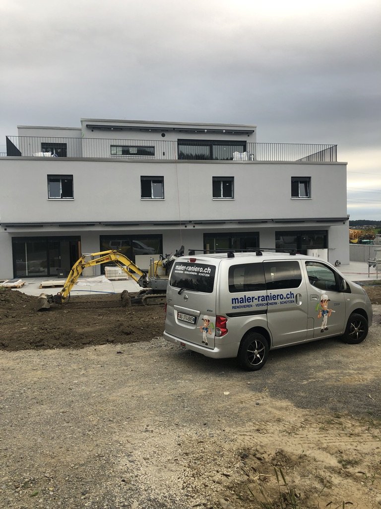 Bauarbeit an modernem weißen Mehrfamilienhaus mit Baumaschine und einem silbernen Firmenwagen auf Baustelle