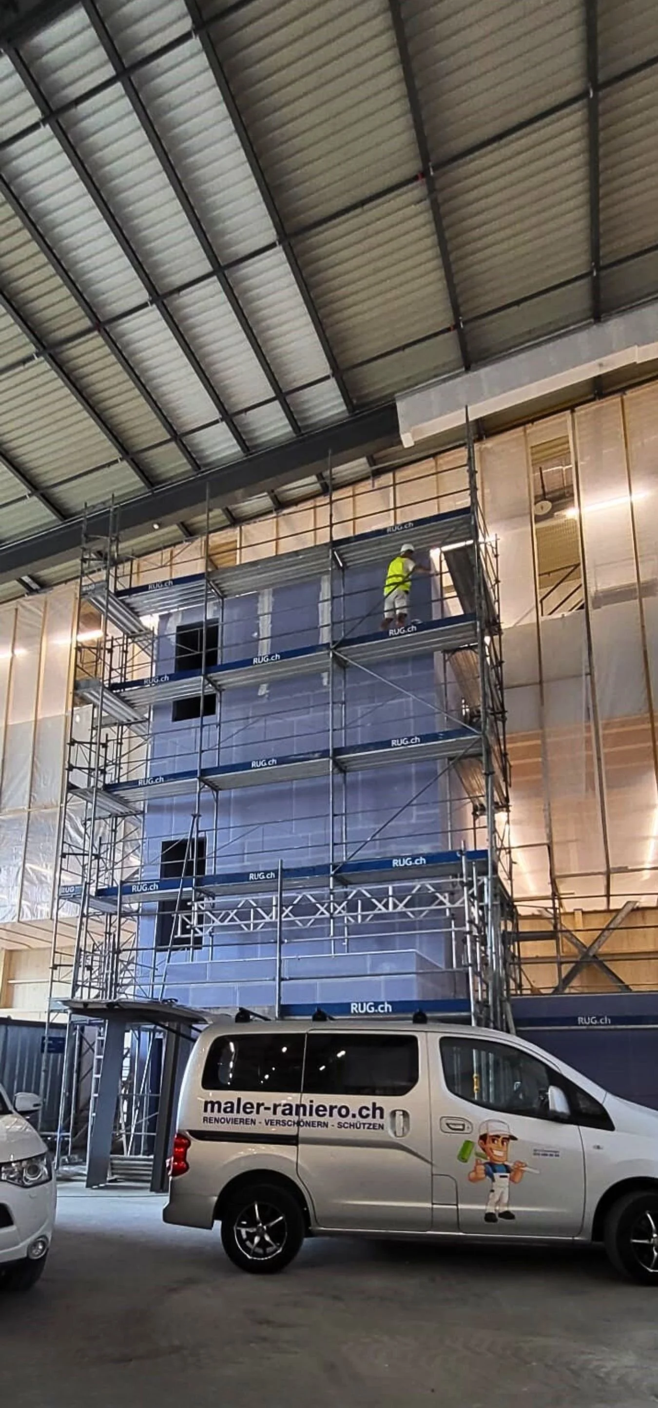 Innenaufnahme eines Gebäudes im Bau mit Gerüsten und Bauarbeitern. Es wird an der Wand gearbeitet, während ein Firmenwagen im Vordergrund steht.