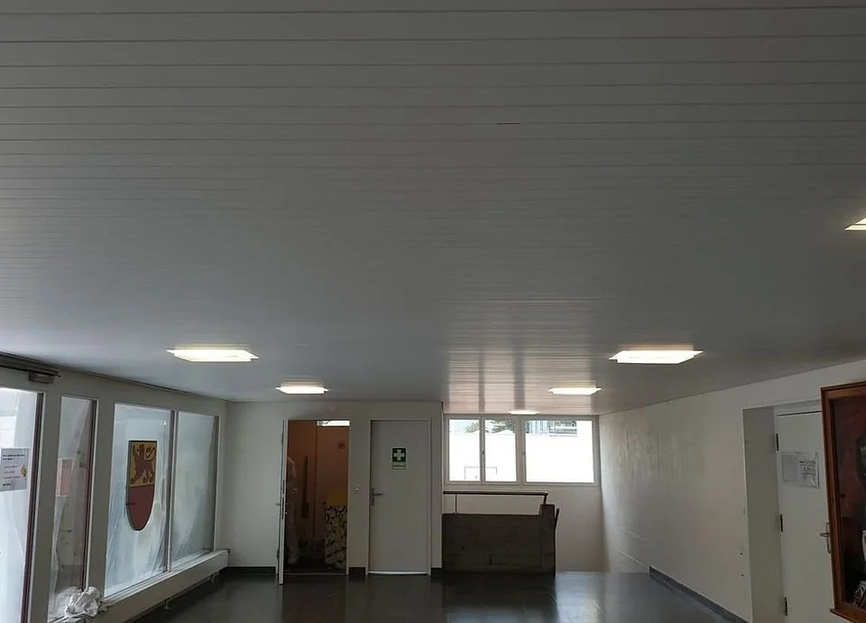 Ein leeres Zimmer mit weißen Wänden, grauem Decke, mehreren Deckenlichtquellen und Fenstern auf der linken Seite sowie einem Fenster in der Mitte.