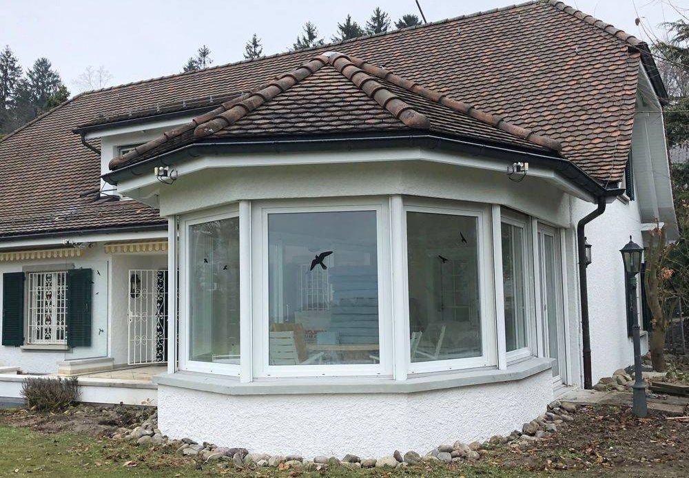 Ein Haus mit weißer Fassade, dunklen Fenstern und einer großen, ovale Veranda aus Glas, umgeben von einer Rasenfläche und Bäumen im Hintergrund.