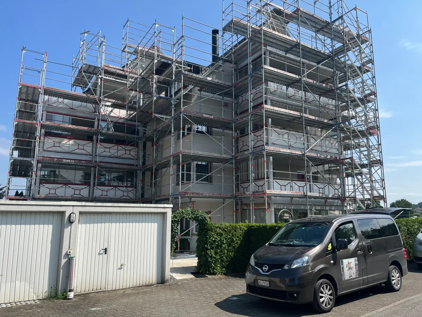 Mehrstöckiges Gebäude unter Baustelle mit Gerüsten, vor einem grünen Busch und einem grauen Fahrzeugparkplatz bei sonnigem Wetter.