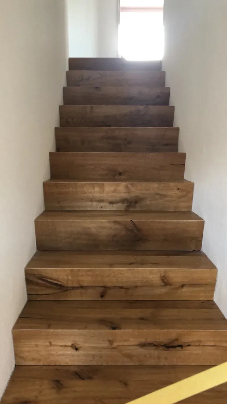 Holztreppe in einem schmalen Flur, führt nach oben, Lichtquelle im Hintergrund.