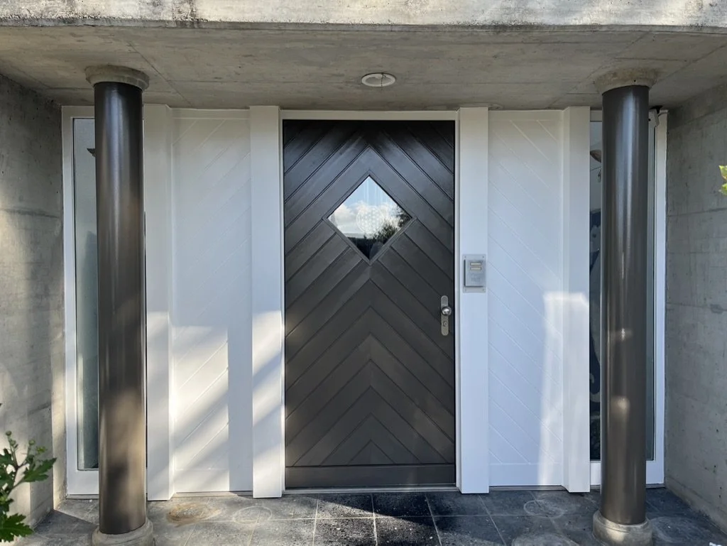Der Eingang zu einem modernen Haus mit schwarzer Holztür mit einem kleinen Fenster in der Mitte, flankiert von weißen Wänden, zwei silbernen Säulen und Seitenfenstern.