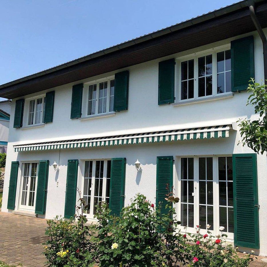 Ein zweistöckiges Haus mit weißen Wänden, grünen Fensterläden, einer gestreiften Markise und Garten mit Rosen.