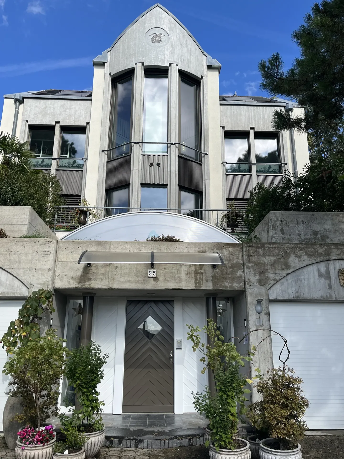 Mehrstöckiges modern gestaltetes Haus mit großen Fenstern, einem weißen und grauen Eingangsbereich, mehreren Pflanzgefäßen mit Bepflanzung und einer blauen Himmel mit wenigen Wolken.