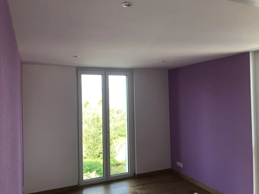 Ein leerer Raum mit weißen und lavendelfarbenen Wänden, Holzboden und großen Fenstern, die einen Blick auf einen grünen Garten bieten.