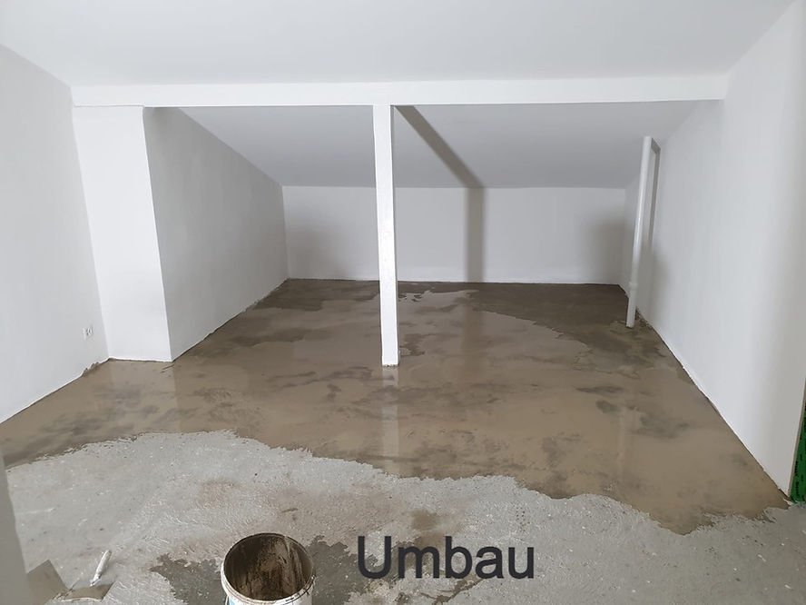 Ein unrenovierter Raum mit unvollständigem Boden und weißen Wänden, teilweise geöffneten Deckenbalken und einer Tür, die zu einem anderen Bereich führt.