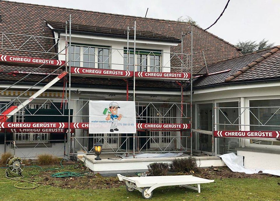 Gerüstsausrüstung vor einem Haus, das renoviert wird, mit einem Banner eines Handwerkers und einer Rasenfläche im Vordergrund.