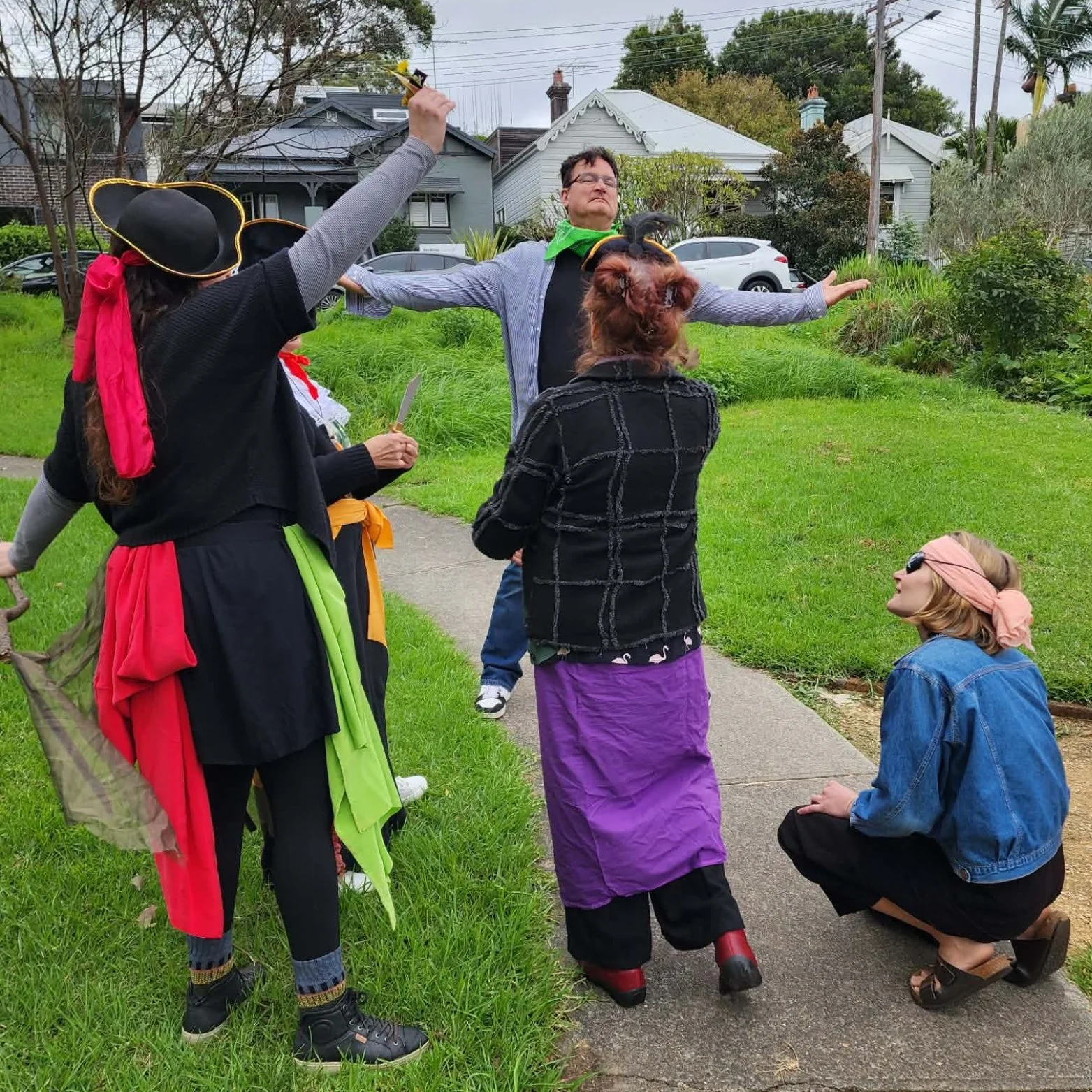 People dressed as pirates and a woman in modern clothing participate in a theatrical or cosplay scene outdoors on a sidewalk near grass and trees.