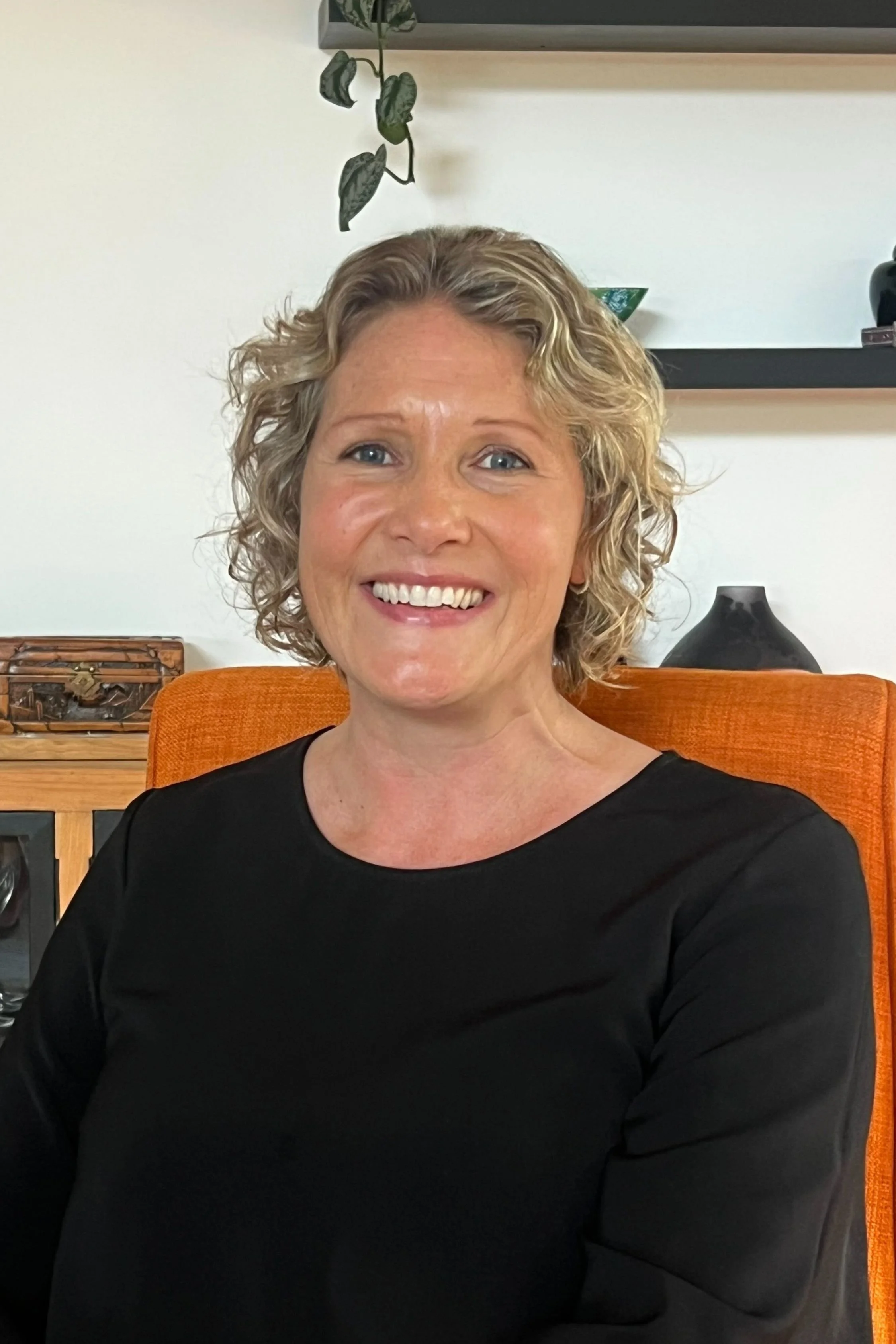 Smiling woman with curly blonde hair wearing a black top seated on an orange chair in a cozy room with shelves and decorative items in the background.