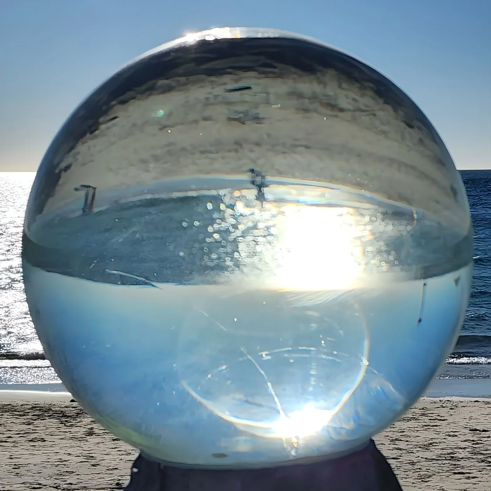 A clear glass ball on a wooden surface with a view of the beach, ocean, and sky reflected inside it, showing a person walking on the beach and the sunlight.