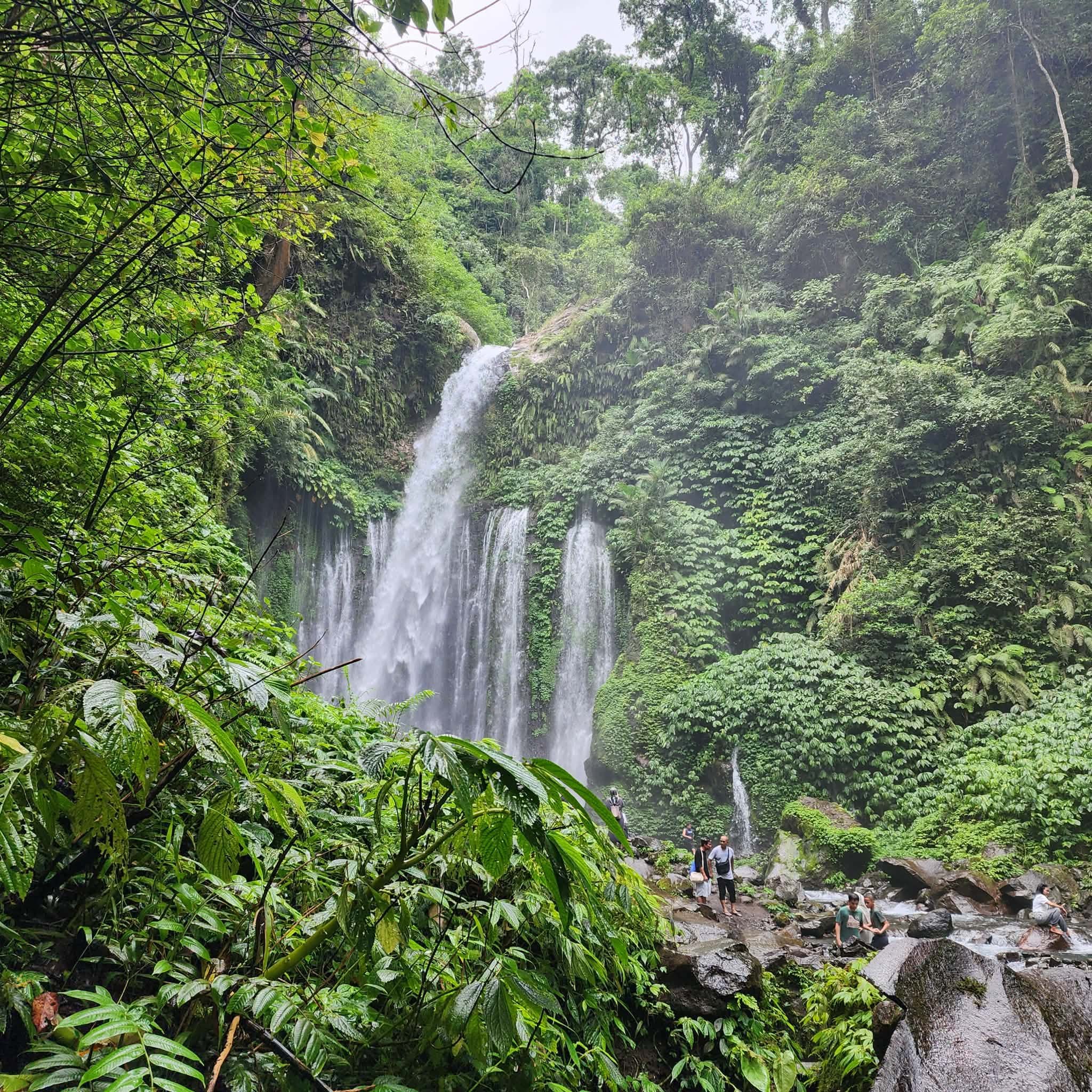 Lush greenery surrounds a waterfall cascading down a rocky cliff into a river, with several people exploring the area.
