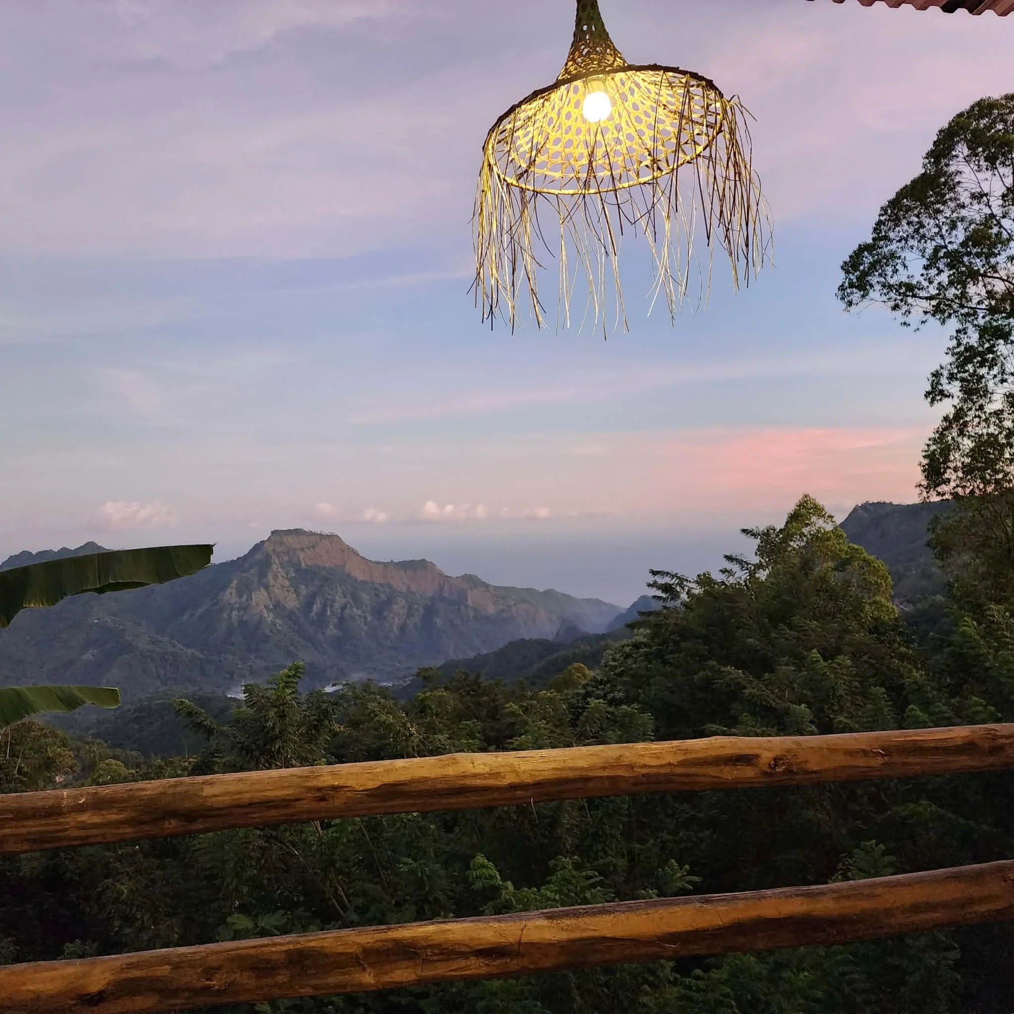 View of mountain range and lush green forest at sunset, with a wooden railing in the foreground and a decorative hanging light fixture overhead.