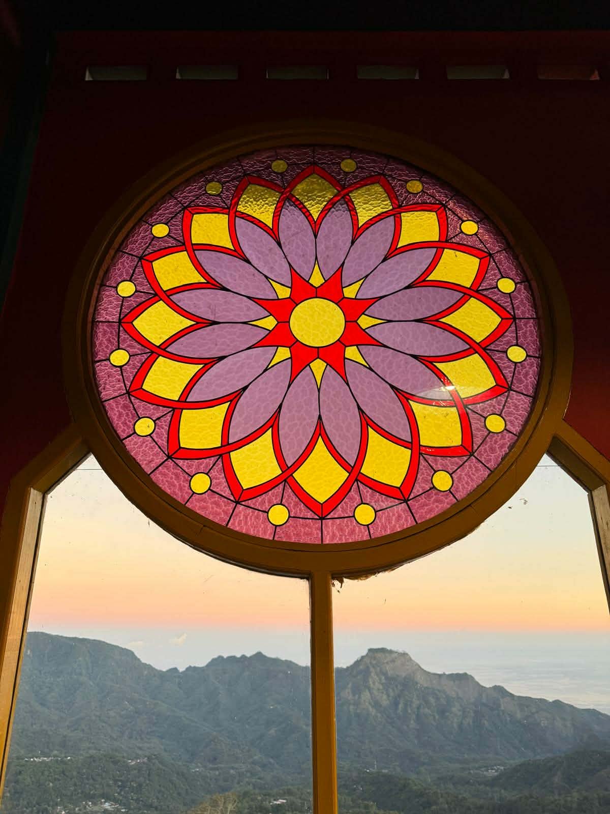 Colorful stained glass window with a floral pattern, overlooking mountains and a sunset sky.
