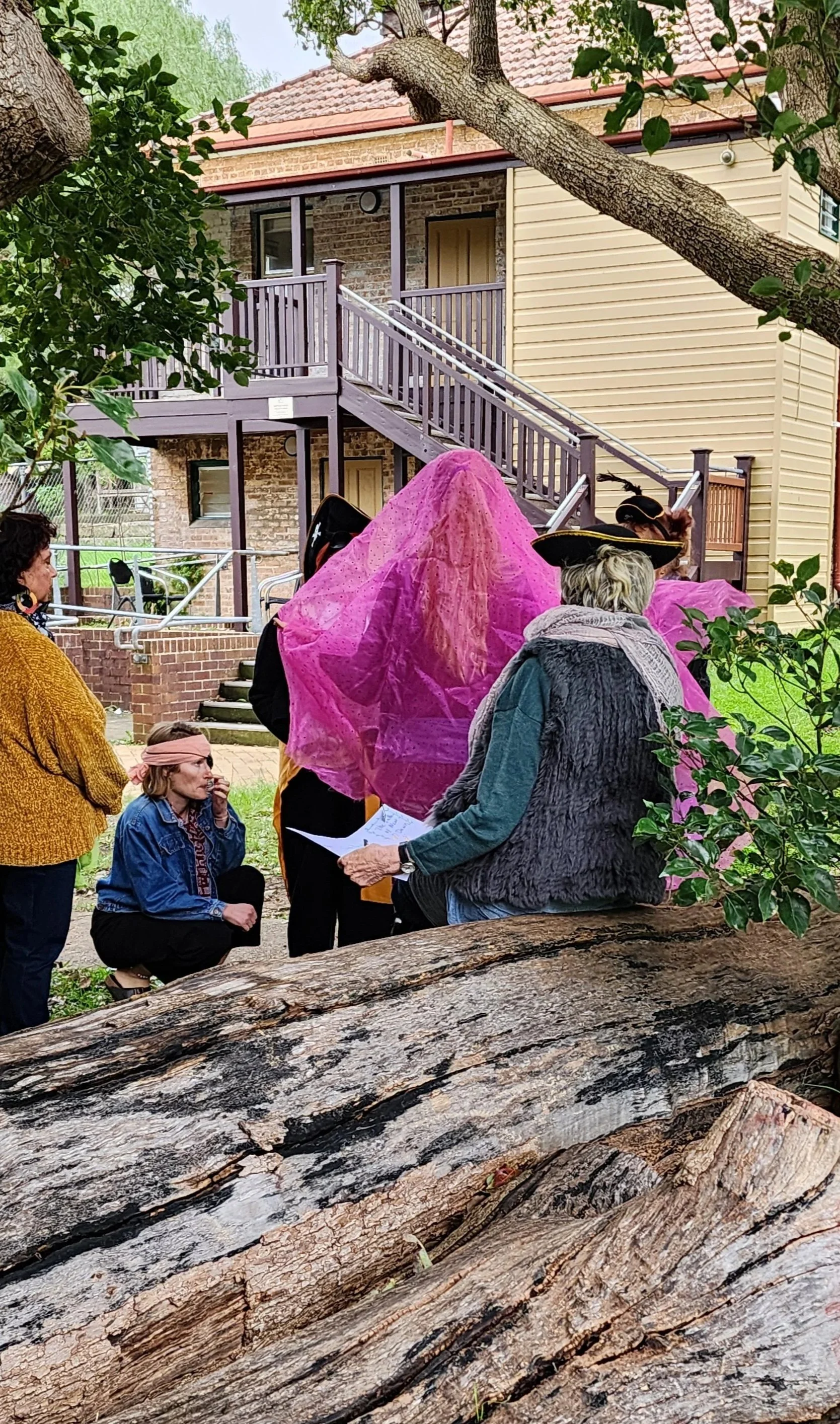 A group of people dressed in costumes, including a woman in pink with a veil and a woman with a pirate hat, gathered outdoors near a large fallen tree log, with a building with a staircase, brick wall, and green foliage in the background.