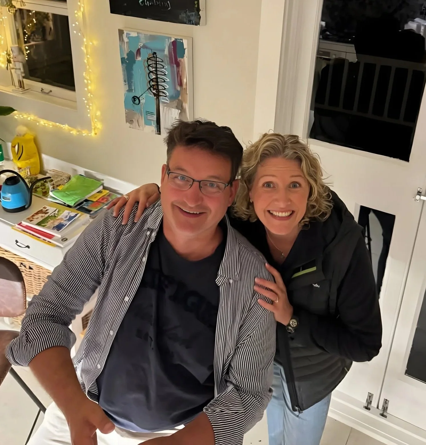 Two people, a man and a woman, smiling and posing together indoors. The man is wearing glasses, a striped shirt over a dark T-shirt, while the woman has curly blonde hair and is wearing a black jacket. The background includes a window with a string of yellow lights, a wall with artwork, and a table with various items and magazines.