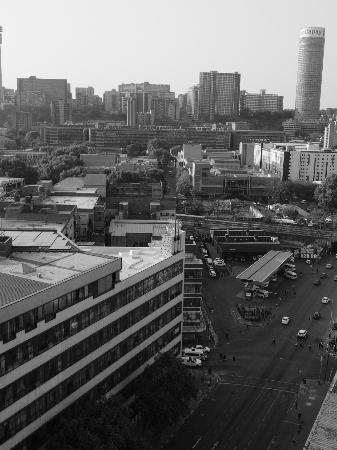 Black and white photo of a cityscape with a mix of tall buildings, trees, and a busy intersection with cars and pedestrians.