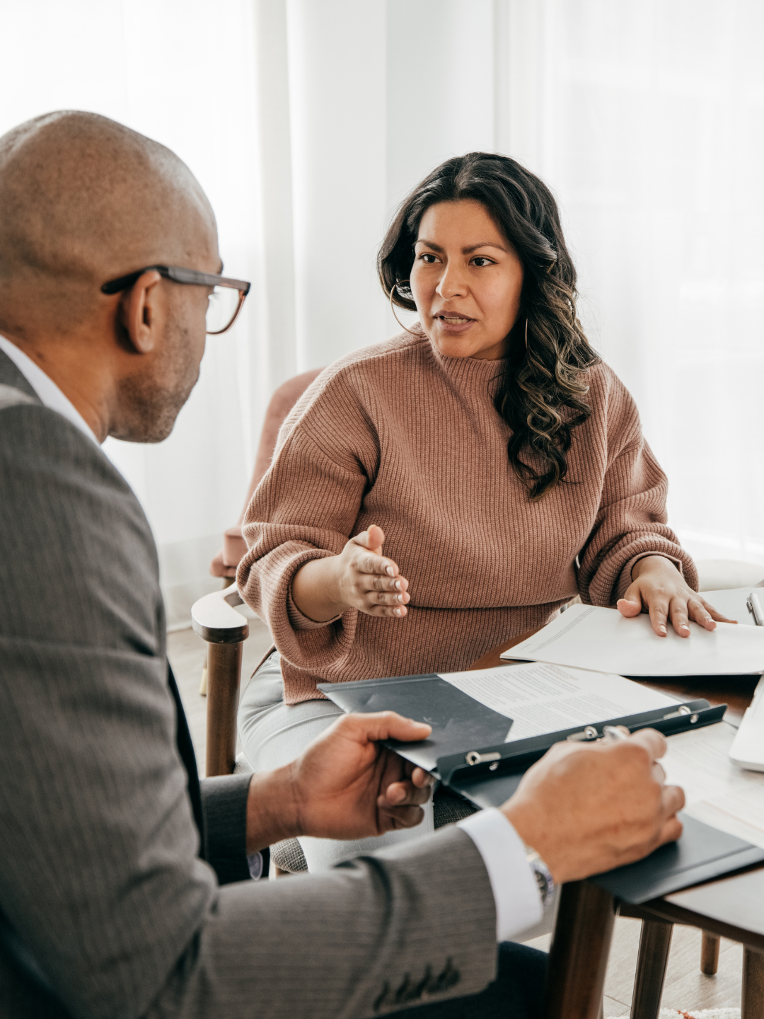 Two people in a meeting, a woman in a brown sweater gesturing with her hand while speaking to a man in a gray suit holding a folder.