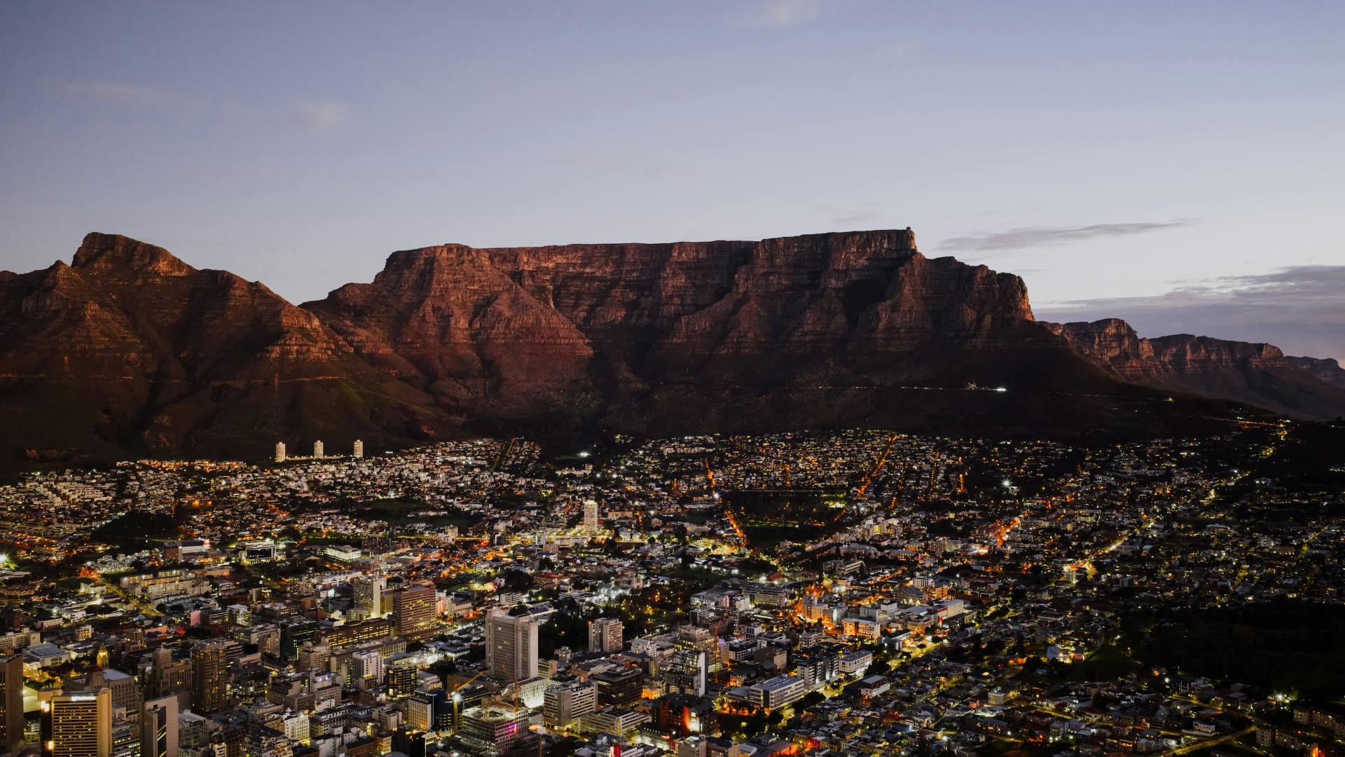 Night cityscape with illuminated buildings in the foreground and mountains in the background under a twilight sky.