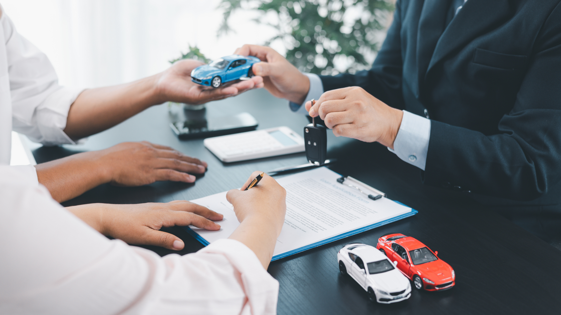 A person in a black suit hands over a blue toy car to another person, while the person in a white shirt writes on a document on a table with two miniature cars, one white and one red, placed nearby.