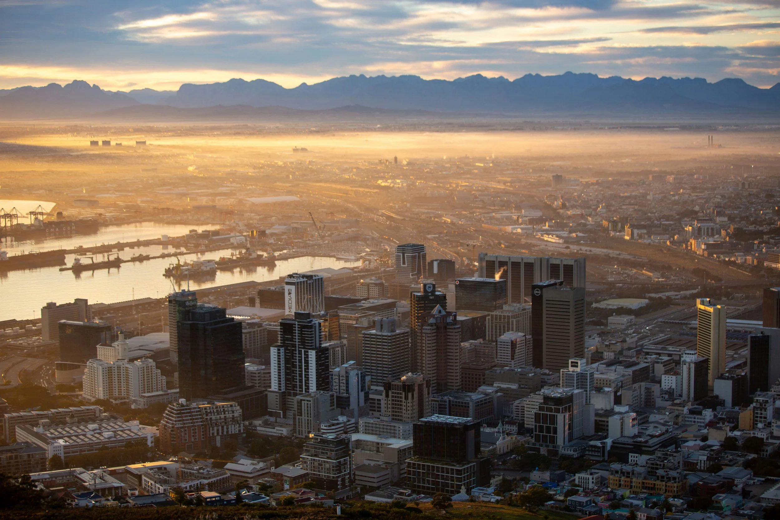 Aerial view of a city skyline during sunrise with mountains in the background, fog over the city, and high-rise buildings in the foreground.
