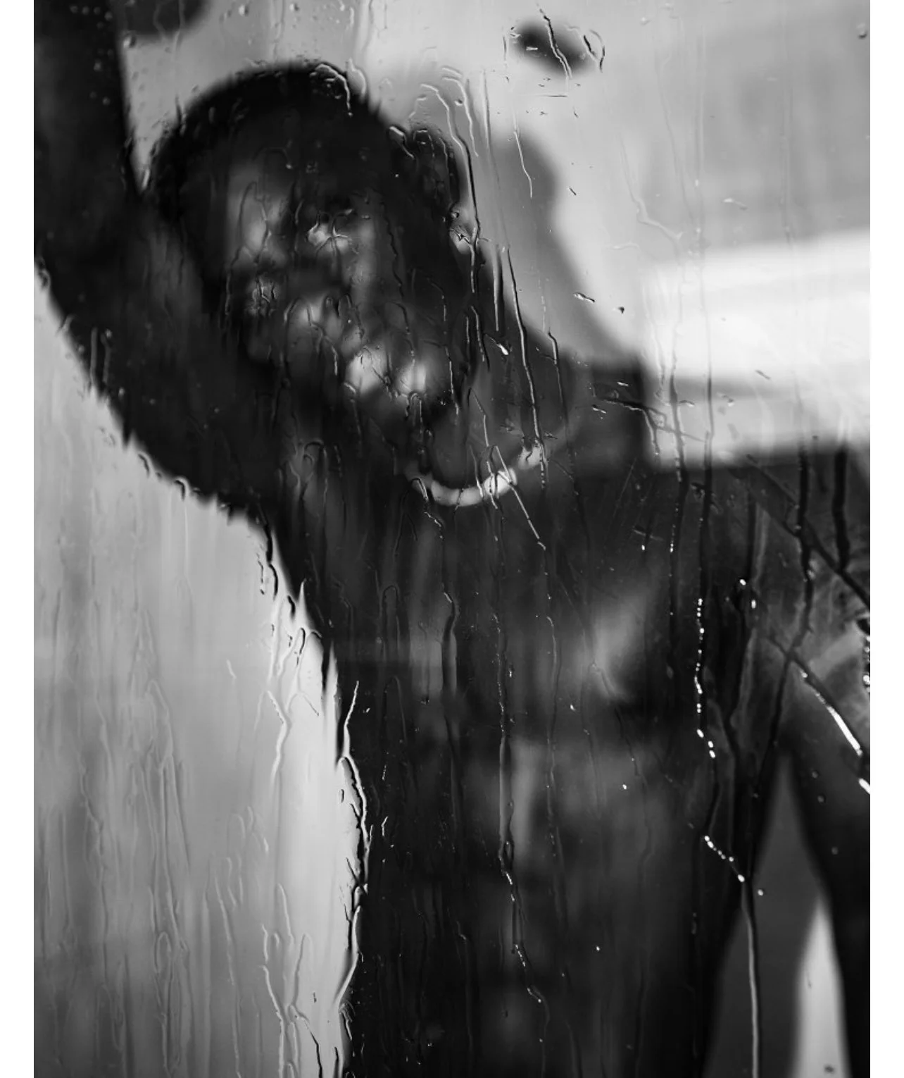 Person with short hair and earrings looking through a rain-covered window, with the reflection of a hand and finger touching the glass.