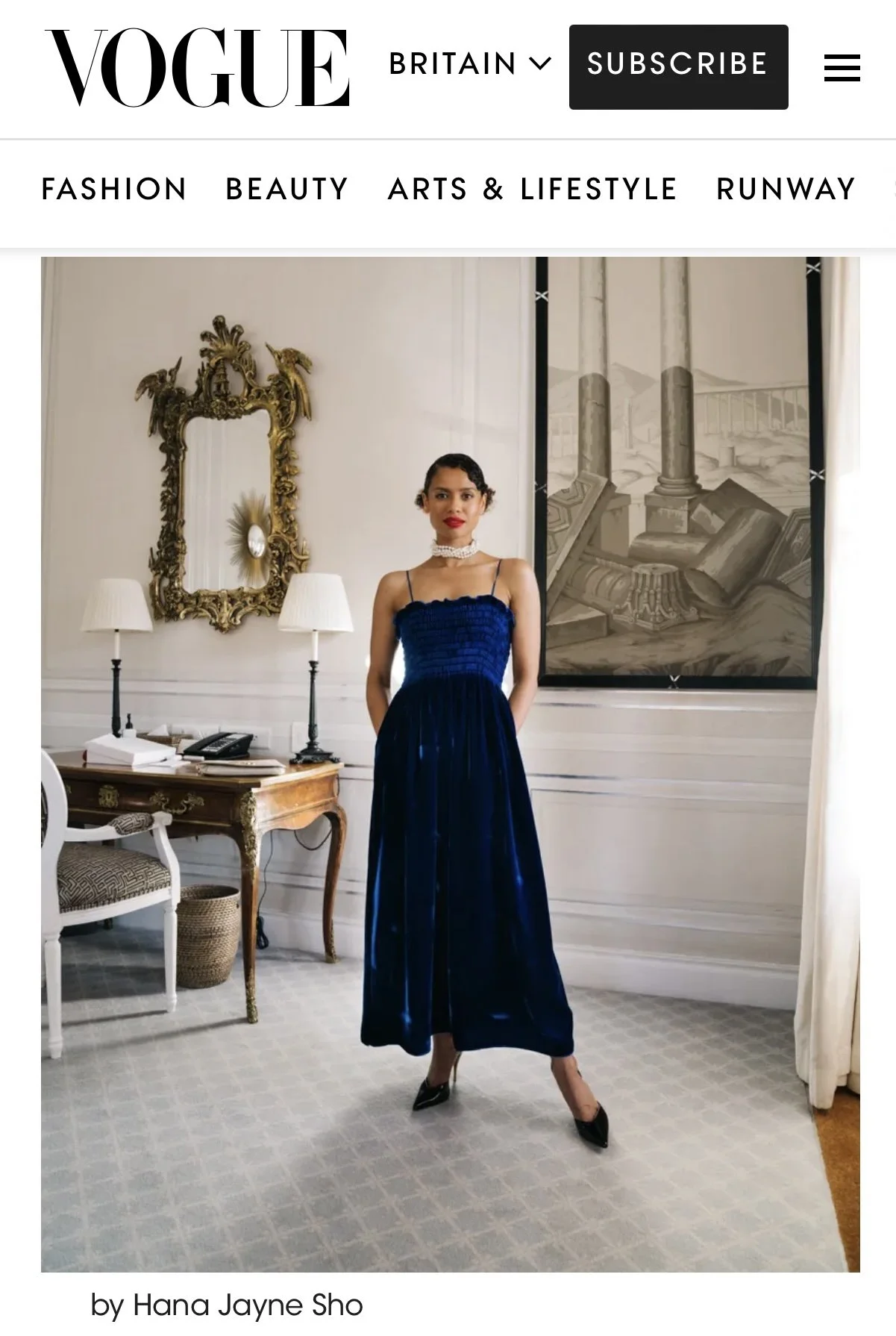 A woman in a strapless blue dress, black high heels, and pearl jewelry standing in an elegant room with a gold ornate mirror, two white lamps, and classical artwork on the wall.