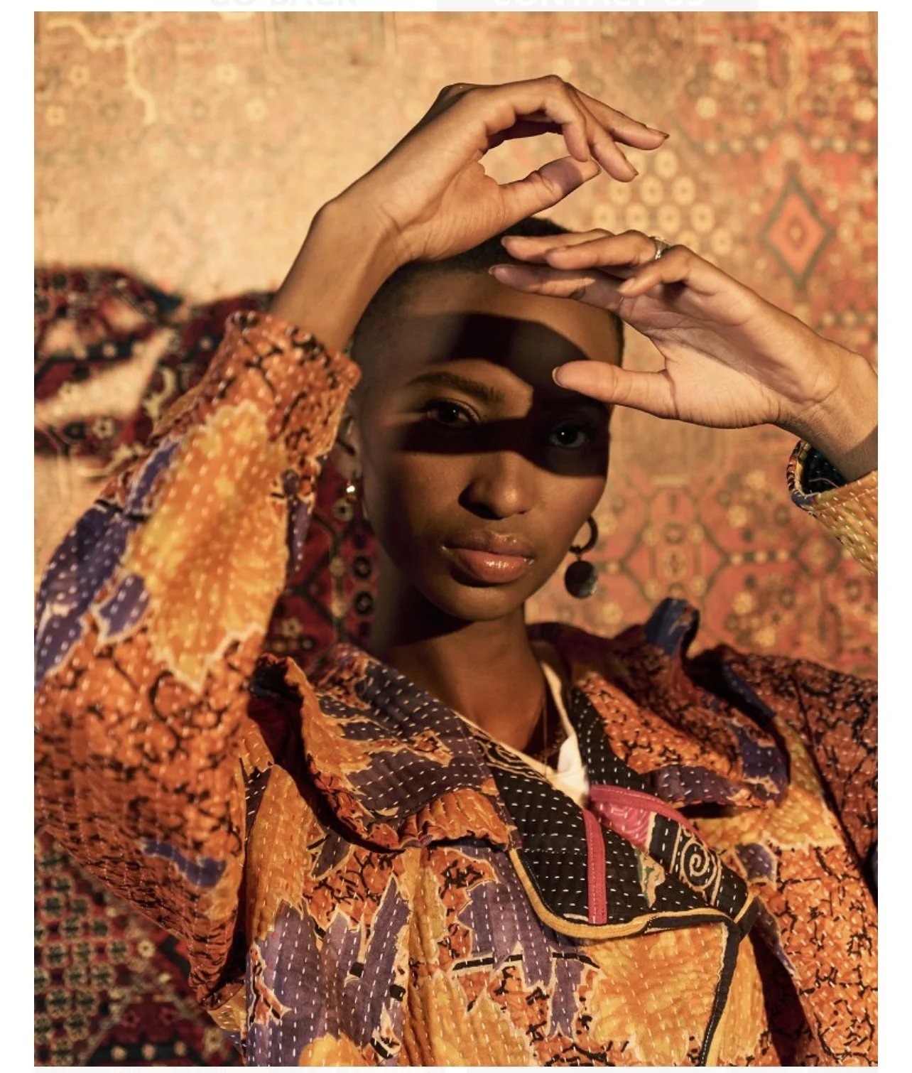 A woman with dark skin and short hair, wearing a patterned orange, purple, and black jacket, holds her hands above her face, creating a shadow with her fingers. She is looking directly at the camera with a neutral expression, in front of a colorful p