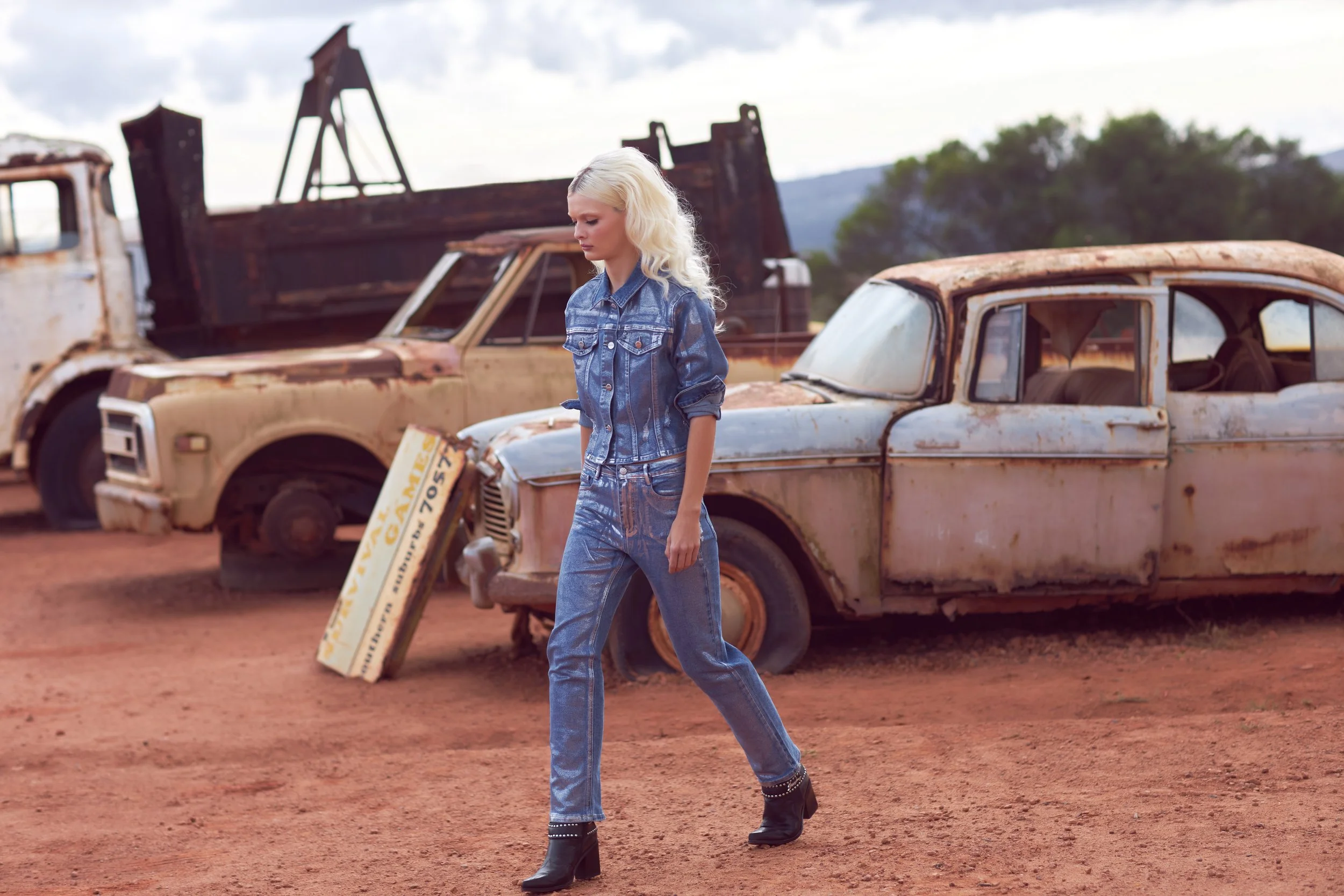 A woman with blonde hair wearing a denim jacket and jeans walking past rusty, vintage cars in a desert-like setting with mountains in the background.