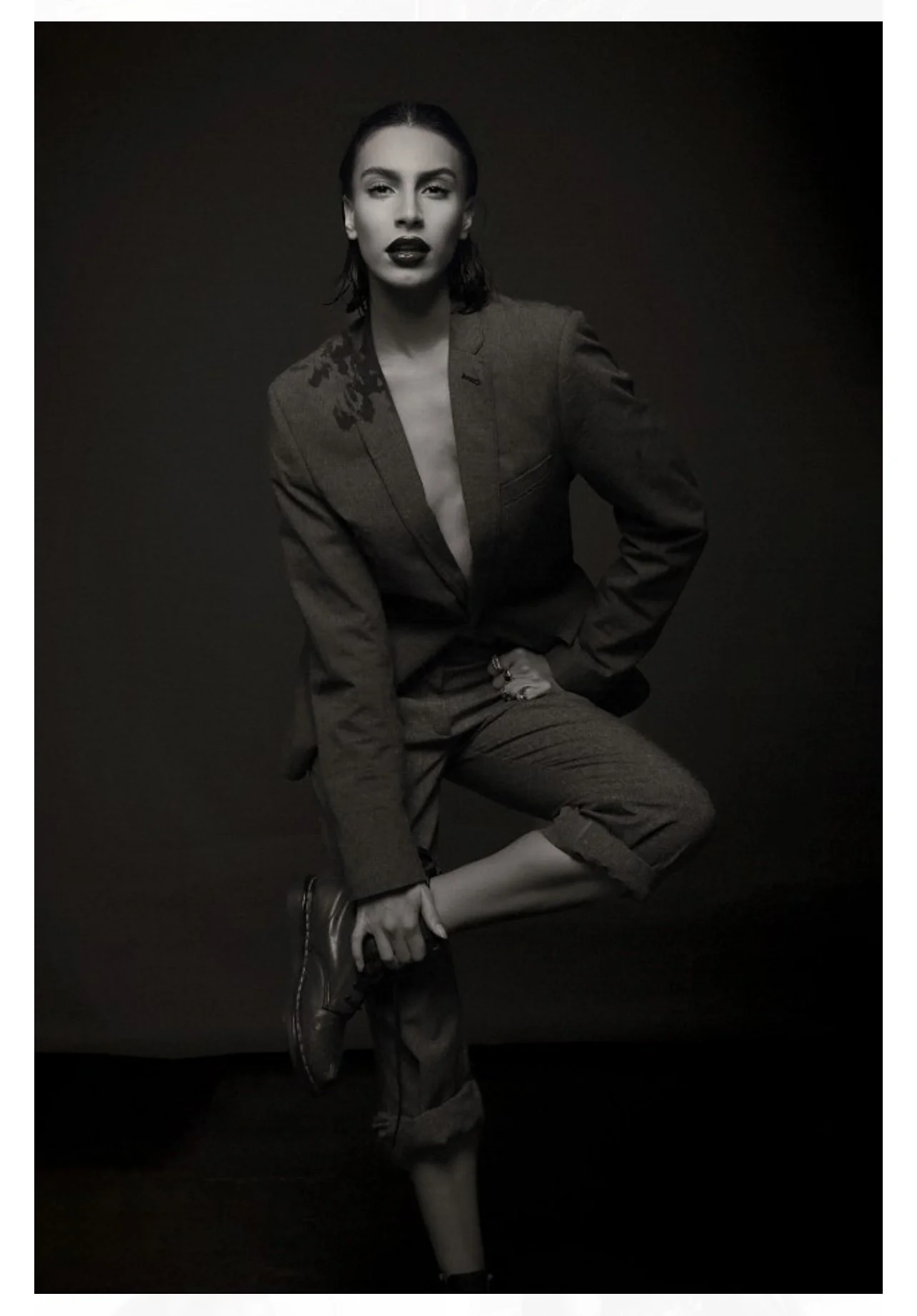 A black-and-white photo of a woman dressed in a blazer and pants, sitting on one knee with her other leg bent, adjusting her sock and shoe.