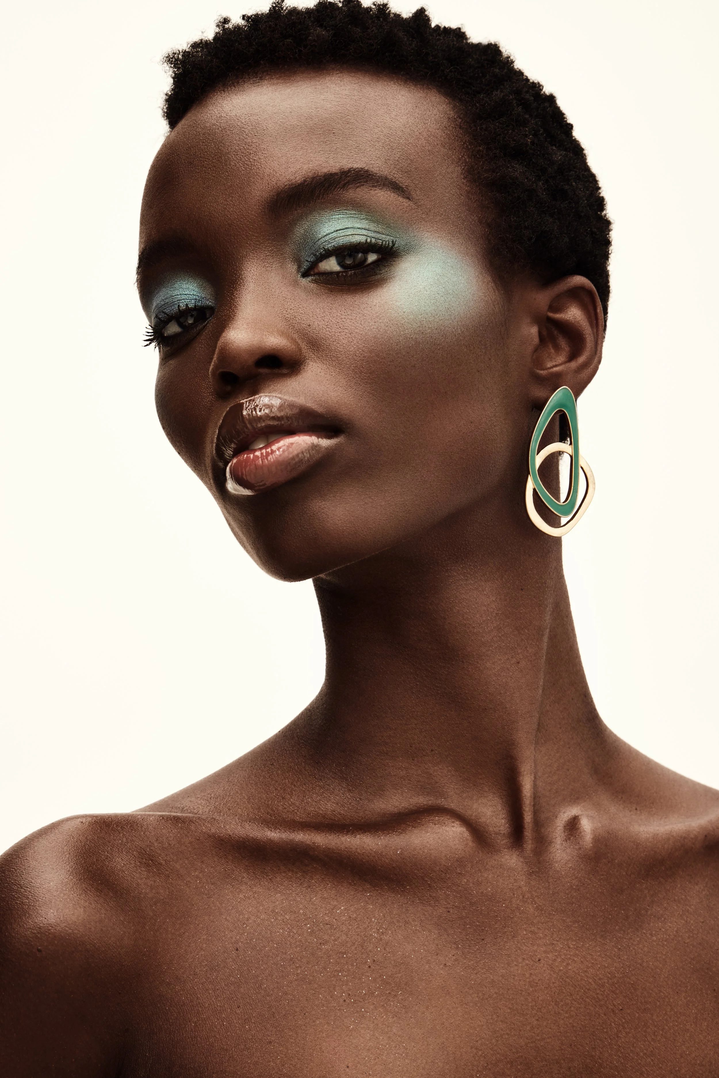 A portrait of a dark-skinned woman with short curly hair, wearing teal and gold earrings, with makeup featuring teal eyeshadow, and a neutral expression against a plain light background.