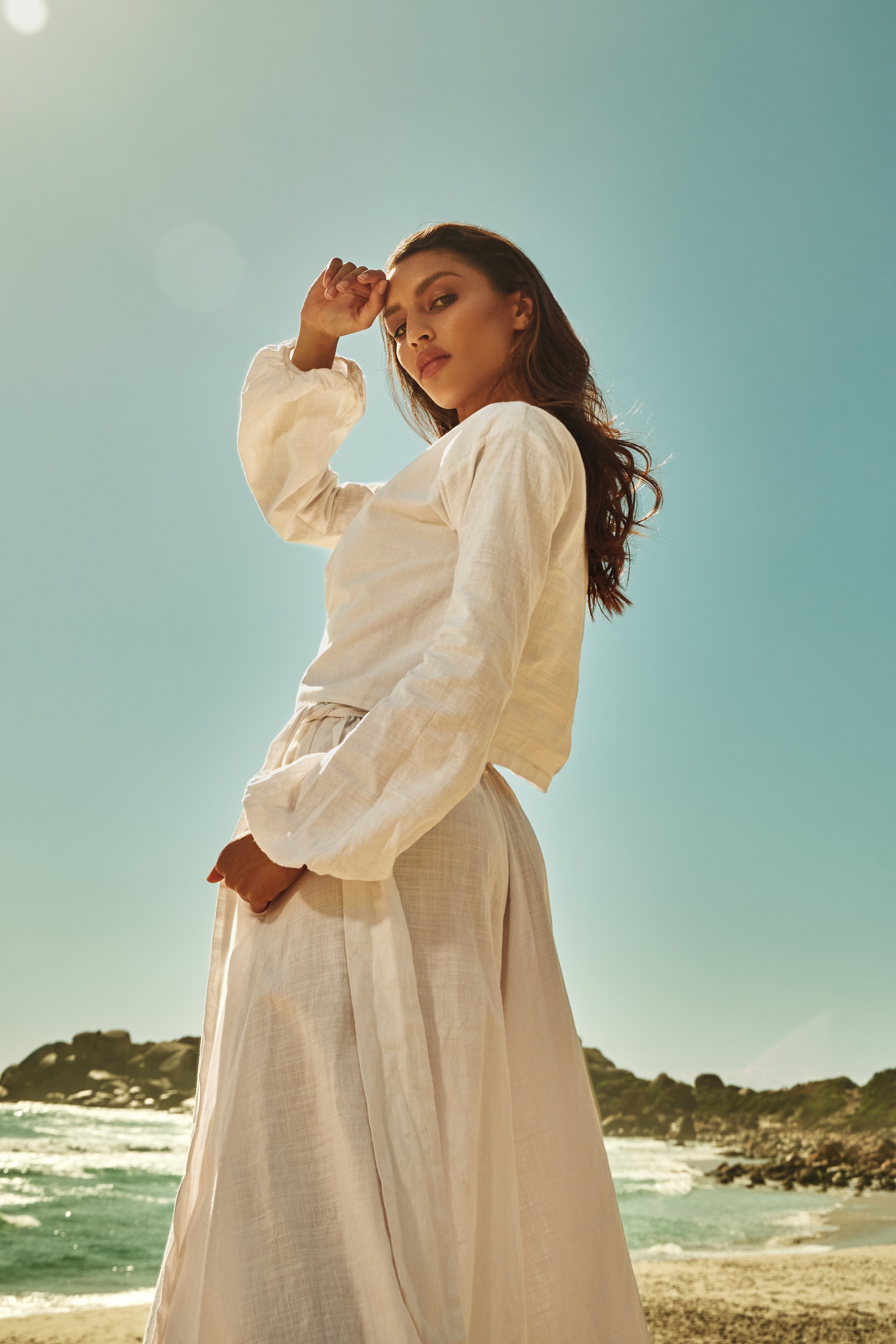 A woman standing on the beach with her hand resting on her head, wearing a white long-sleeve shirt and a flowing beige skirt, with the ocean and rock formations in the background under a clear blue sky.