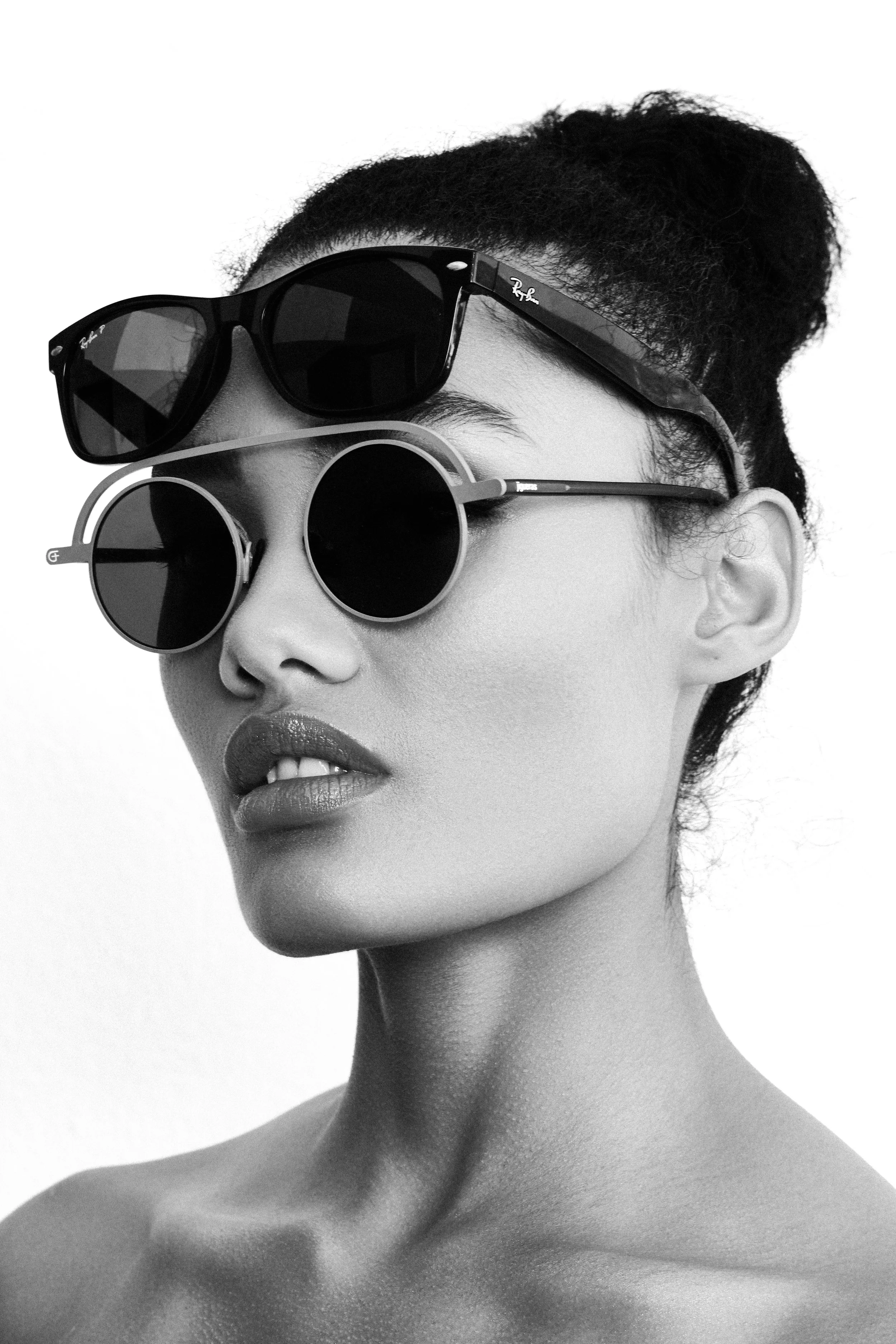 Black and white photo of a woman with sunglasses, with one pair on top of her head and the other over her eyes, looking slightly to the side.