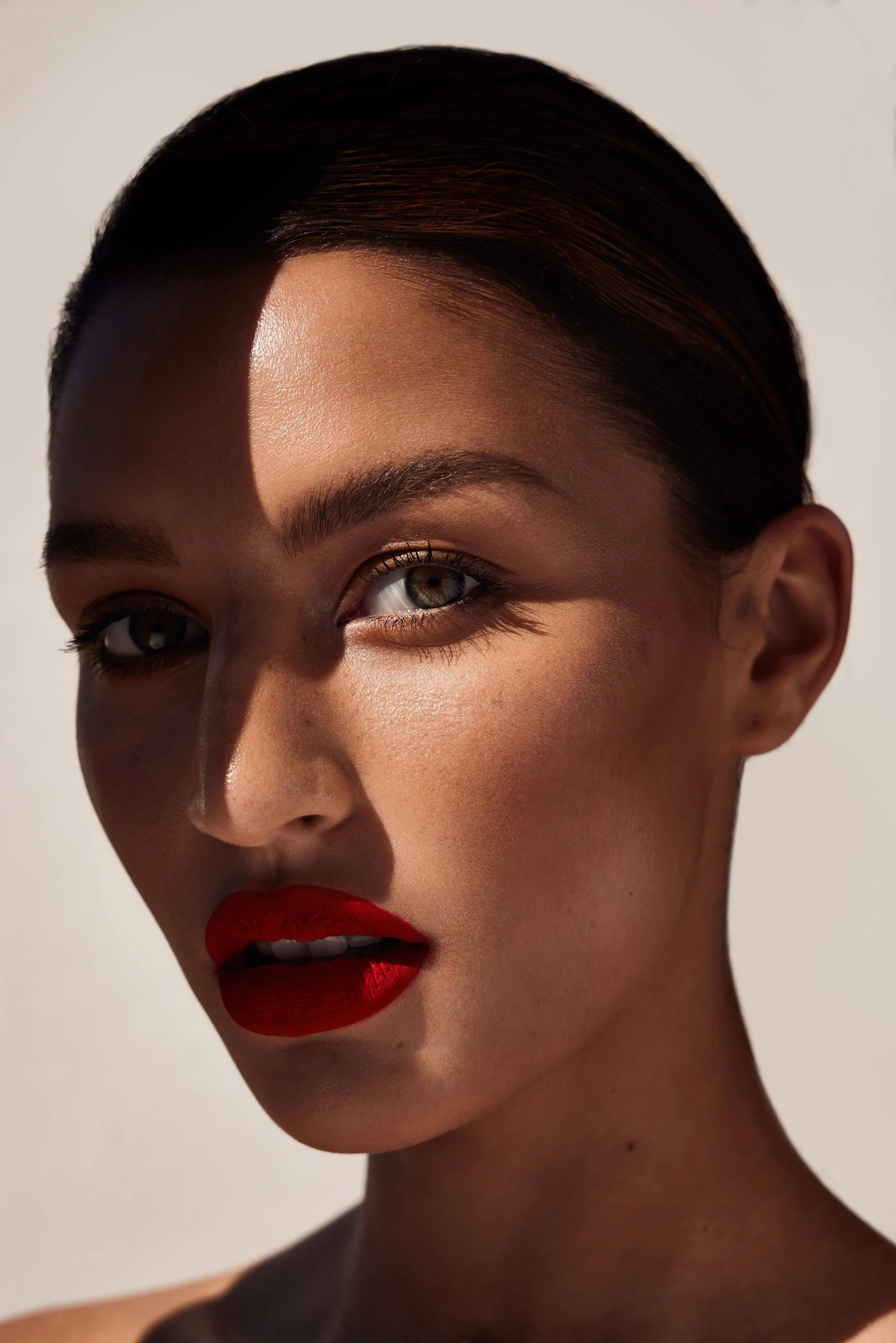 Close-up of a woman with slicked-back hair, wearing red lipstick, with sunlight creating a shadow across her face.