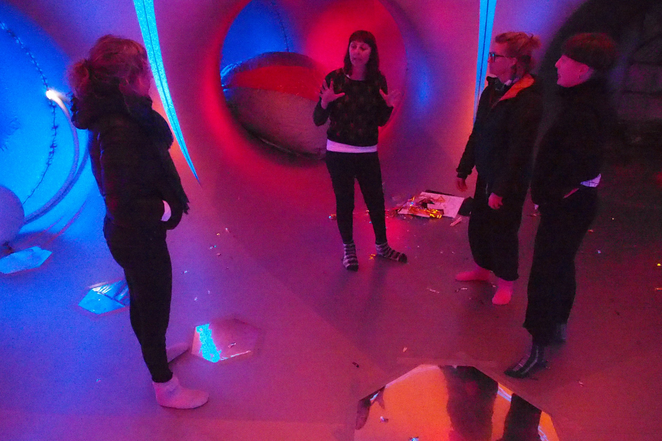Four women in a dimly lit, colorful room with vibrant, neon lighting. One woman is standing in the middle, gesturing with her hands, possibly explaining something, while the others listen. The background has a large circular, reflective or tunnel-like structure, and the floor has scattered items and reflective surfaces.